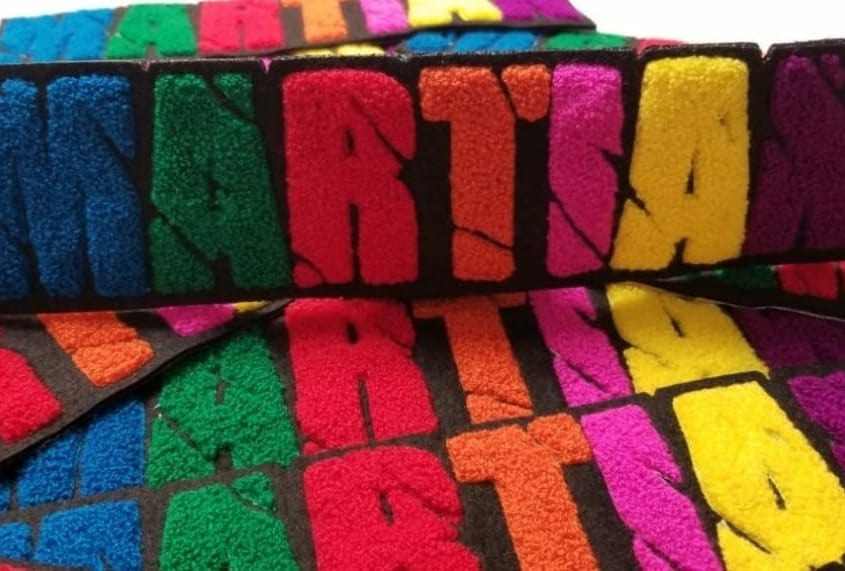 Bulk chenille felt patches in multi color spread that says ‘MARTIAN’ in caps lock capital English alphabet. M in blue yarn, A in green, R in red, T in orange, I in pink, A in yellow and N in fluffy purple on the black scrim felt and twill. The script letters show cut off scars passing through entire wording.