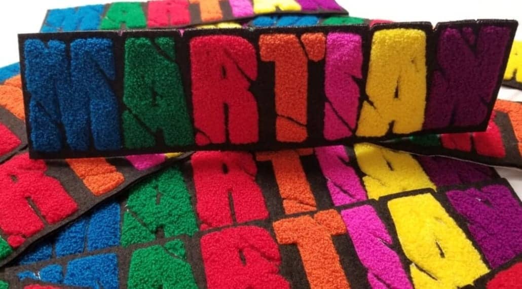 Bulk chenille felt patches in multi color spread that says ‘MARTIAN’ in caps lock capital English alphabet. M in blue yarn, A in green, R in red, T in orange, I in pink, A in yellow and N in fluffy purple on the black scrim felt and twill. The script letters show cut off scars passing through entire wording.