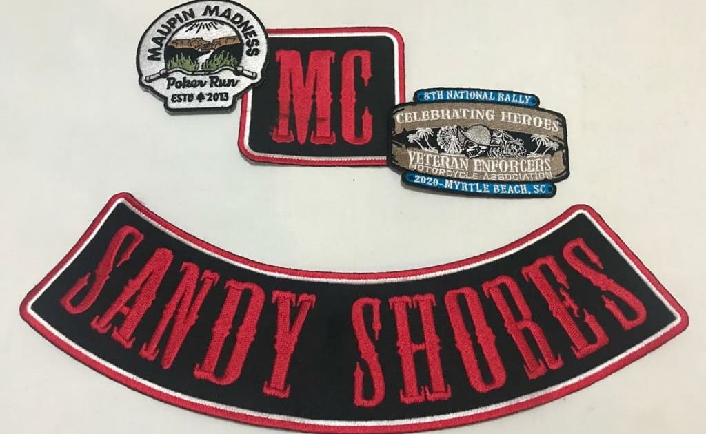 a big banner shows set of custom made embroidery patches for bikers. A black fabric embroidered rocker in the bottom with red thread that says ‘SANDY SHORES’ having white and red outline embroidery. A custom shape horizontal shape in memory biker patch for 8TH NATIONAL RALLY VETERAN ENFORCERS MOTOCYCLE ASSOCIATION of the left top. A square biker MC patch pairing features of large size rocke in red and white embroidery border on black fabric and letters in dense red thread in the center. The fourth biker rally patch on the right top represents ‘MAUPIN MADNESS’ on the top, sceneries and vallies in the center over the handle of bike and ‘POKER RUN’ emrbroidered in the bottom EST 2013.